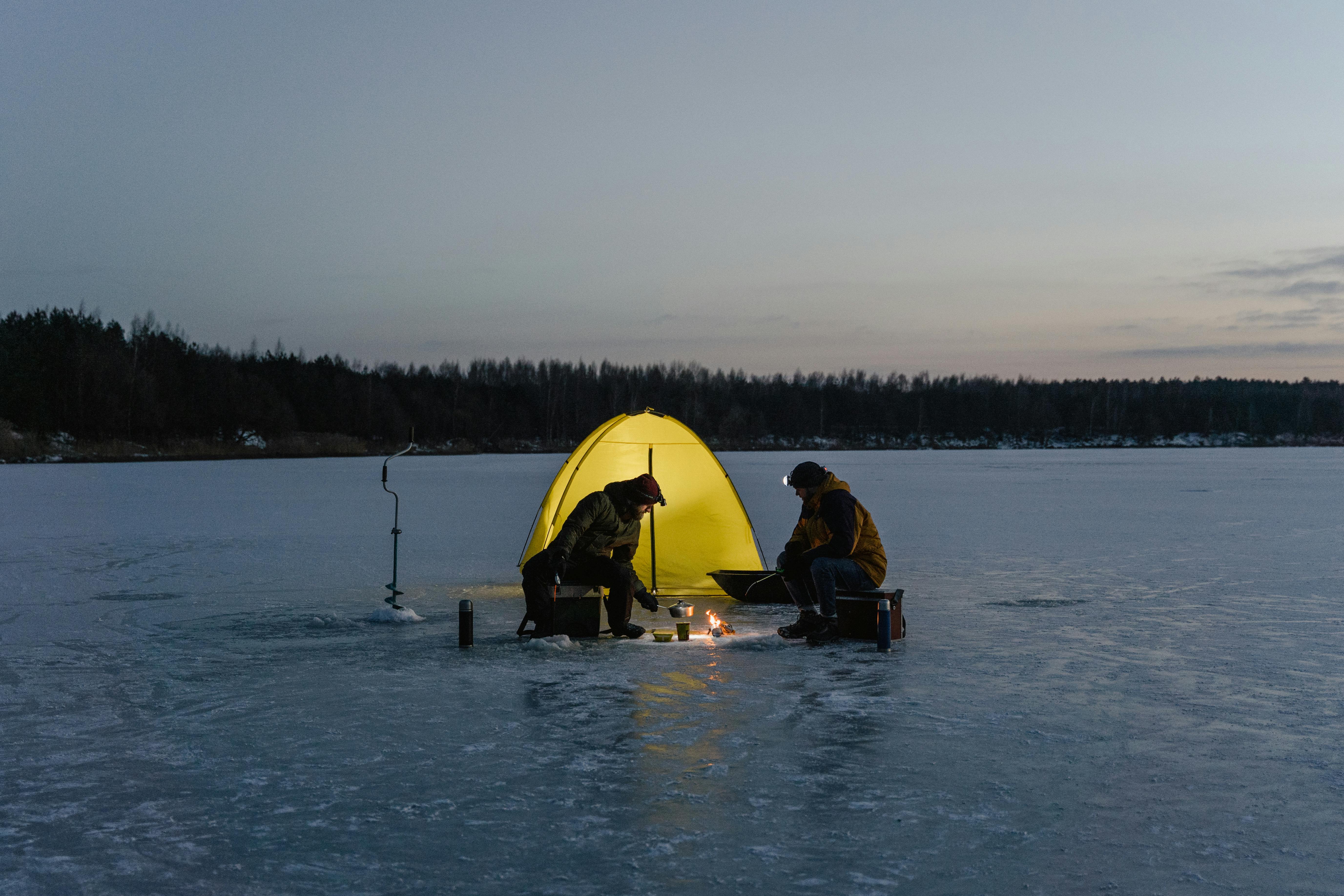 Two people ice fishing under a yellow tent on a frozen lake at night.